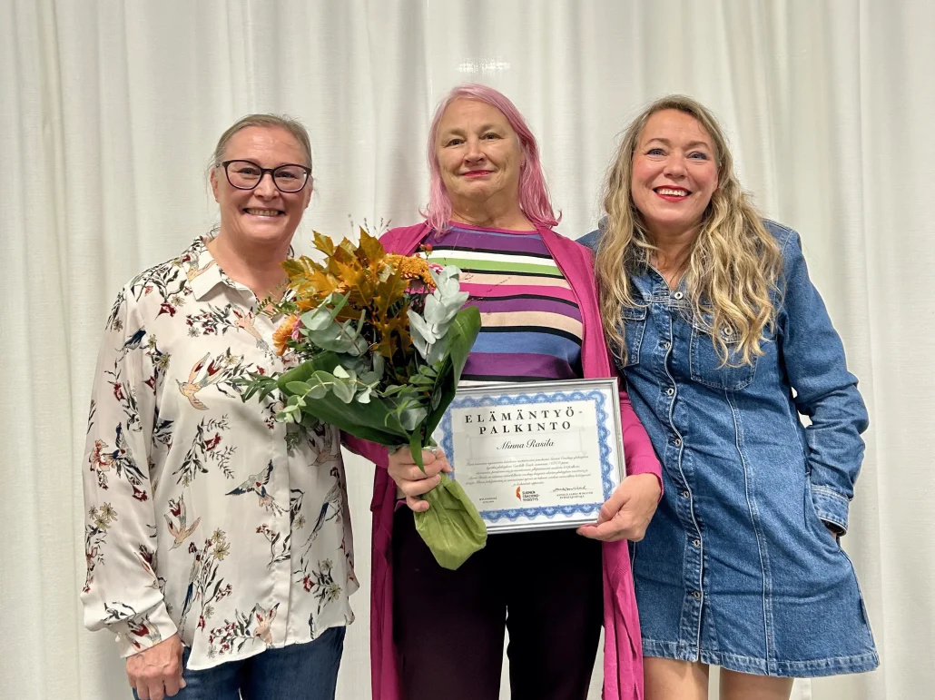 Yhteiskuvassa palkinnon saaja Minna Rasila, jolla on kukkakimppu ja elämäntyöpalkinnon diplomi. Vierellä vasemmalla Suomen Coaching-yhdistyksen puheenjohtaja Annele Aarni-Wiklund ja oikealla varapuheenjohtaja Anna Noramaa.